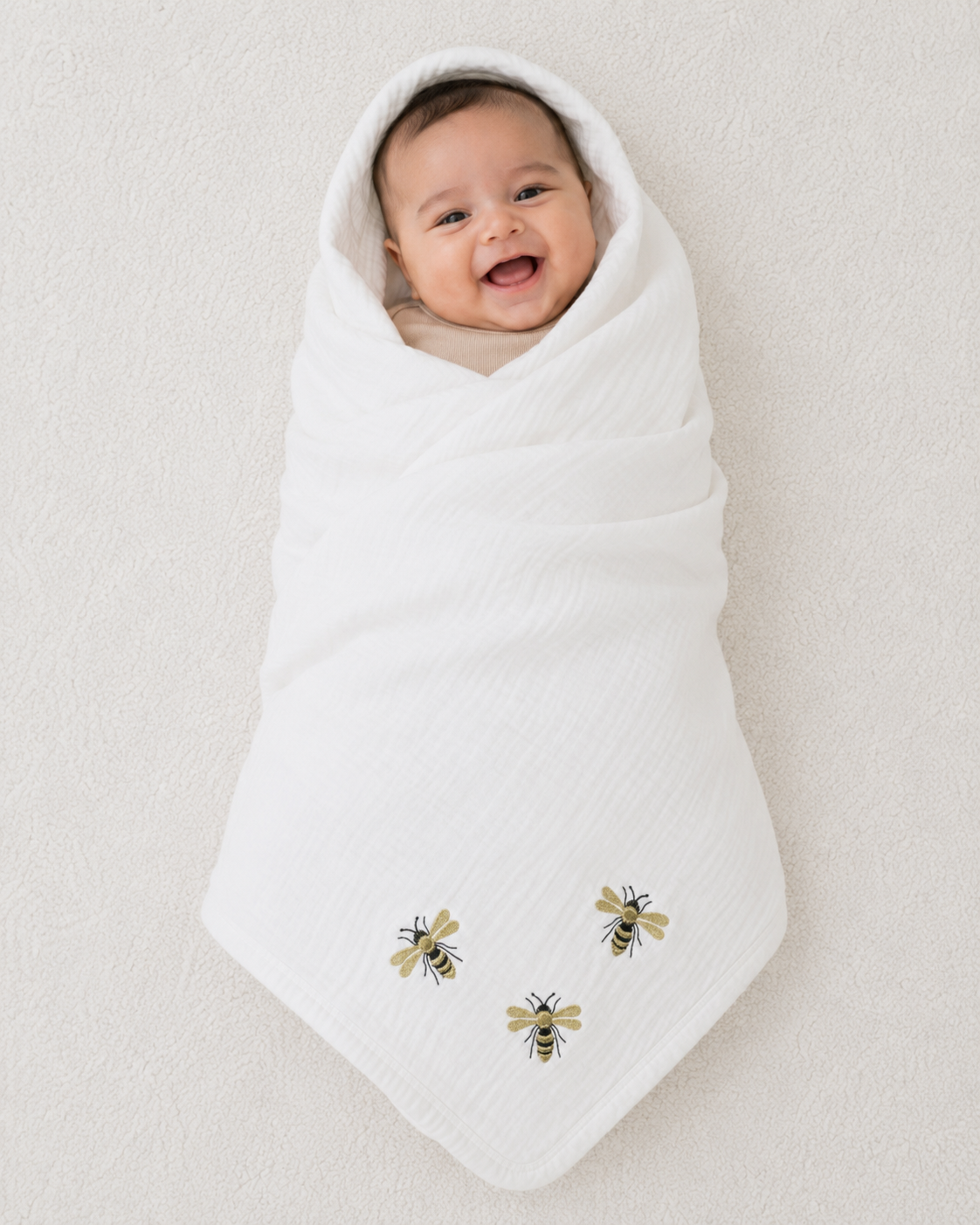 Smiling baby swaddled in soft white muslin blanket with gold embroidered bee design