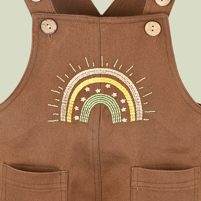 Brown baby dungarees with rainbow embroidery detail.