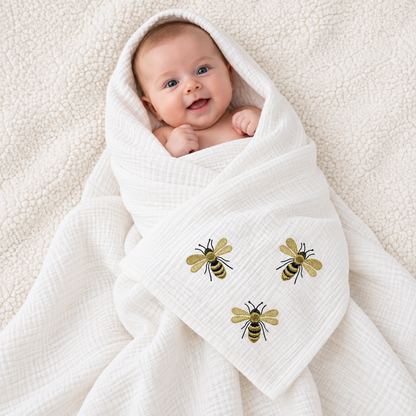 Baby wrapped in a white muslin blanket with three gold and black embroidered bumblebee details.